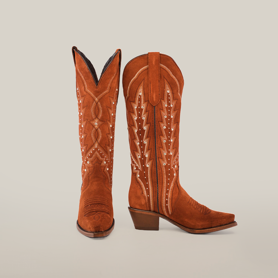 The Caroly Boot, now known as the Caroly Brick Suede, showcases brown cowboy boots with intricate white stitching and decorative designs. Featuring a tall shaft, slightly angled heel, and snip toe against a plain white background.