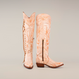 The Emma Alaska Orix-Tall Shaft boots are handcrafted tall tan leather cowboy boots with a subtle antler pattern, featuring a side zipper and heel on one upright boot and decorative lacing on the other against a plain white backdrop.