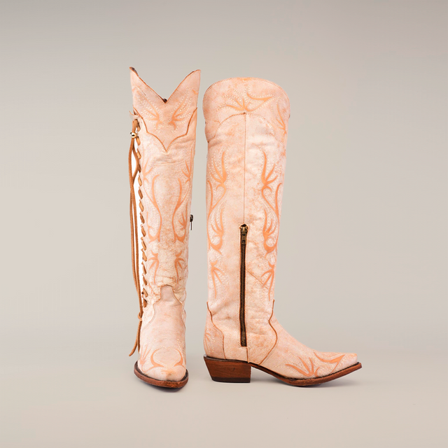 The Emma Alaska Orix-Tall Shaft boots are handcrafted tall tan leather cowboy boots with a subtle antler pattern, featuring a side zipper and heel on one upright boot and decorative lacing on the other against a plain white backdrop.