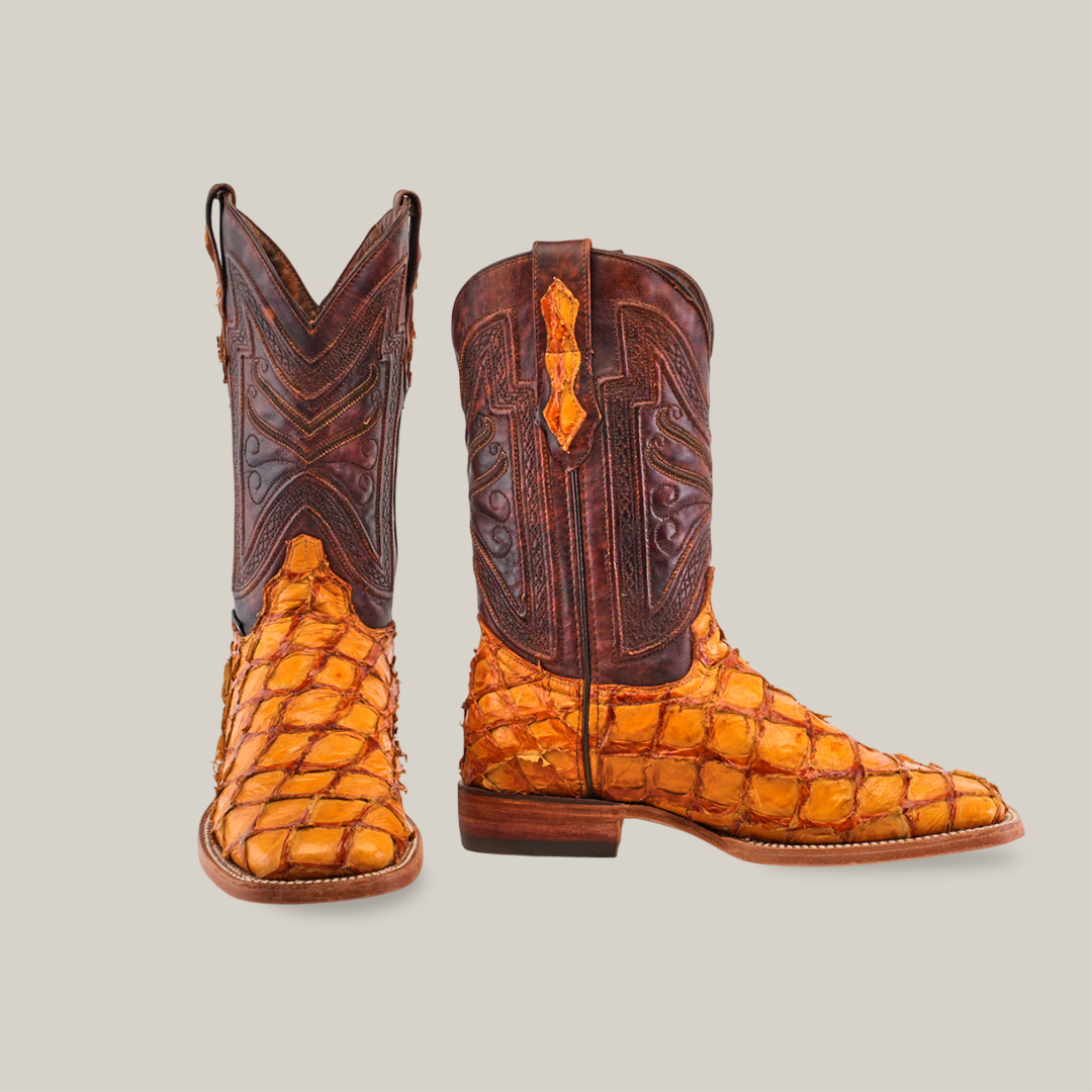 The Exotic Piraruco Fish - Brandy boots feature a square toe in orange-toned pirarucu leather with a scale-like texture. Their dark brown shafts showcase ornate stitching set against a plain, beige backdrop.