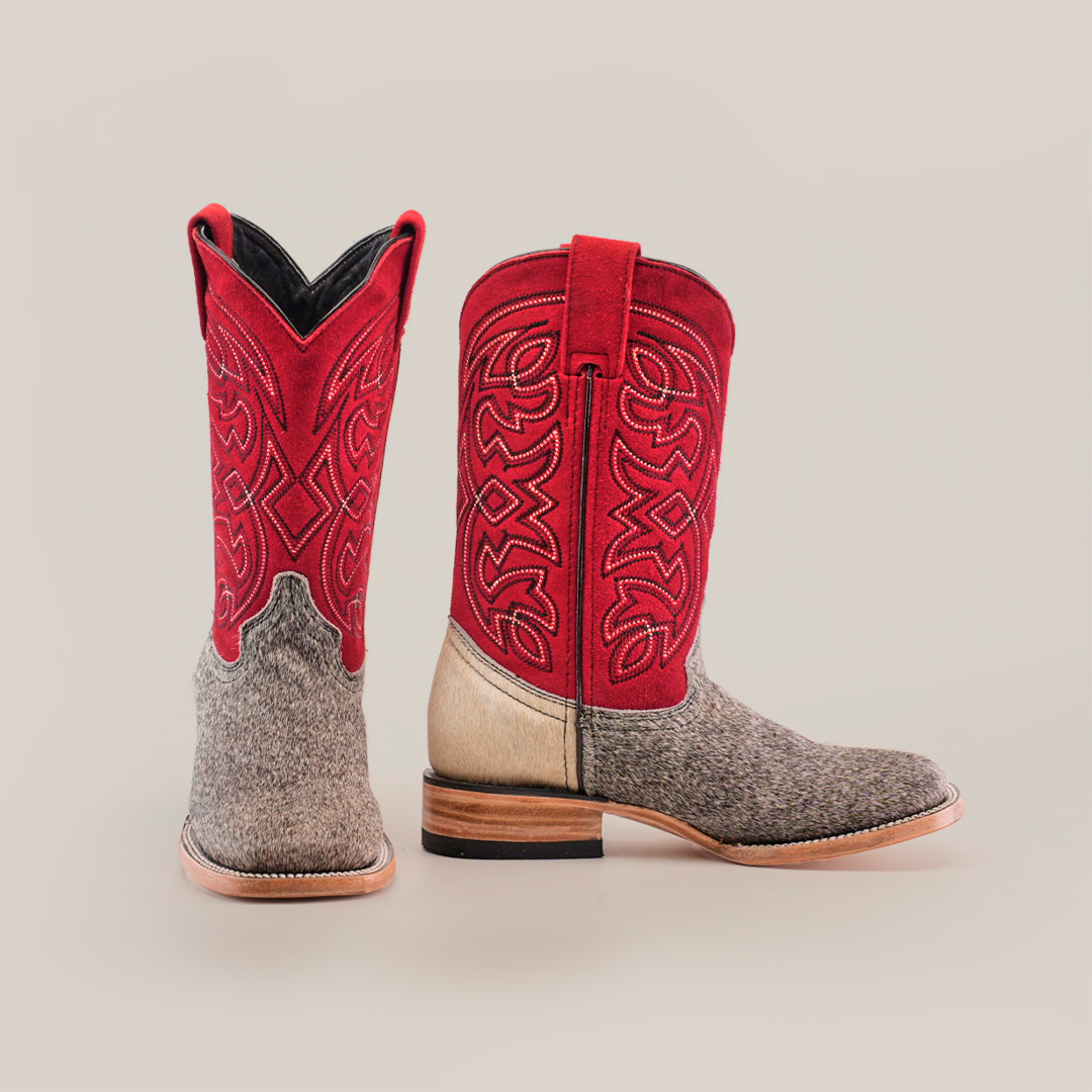 The Cowhide Hair Pinto Square Toe boots feature red intricately stitched shafts, gray textured hair-on-hide leather on the foot and heel, cowhide material, wooden soles, and a square toe. They are displayed upright with one facing forward and the other to the side.