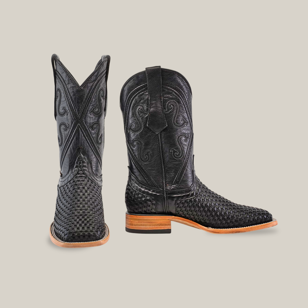 The Texas Basket Weave Black boots showcase a premium cowhide leather with intricate patterns on the toe and shaft. They feature a square toe, wooden heels, decorative stitching, with one standing upright and one resting sideways against a white background.